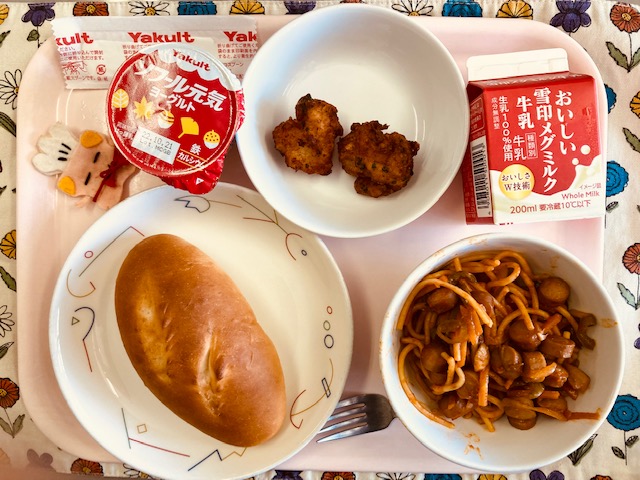 ピンクのトレーに給食が右上から反時計回りで牛乳、魚のふわっとあげ、ヨーグルト、コック帽をかぶった布製マスコット、ミルクパン、スパゲッティナポリタンと並べられている写真