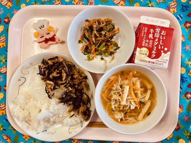 ピンクのトレーに給食が右上から反時計回りで牛乳、小松菜のえのき和え、コック帽をかぶった布製マスコット、ぶたにく丼、そうめん汁と並べられている写真