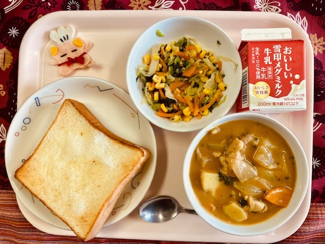 ピンクのトレーに給食が右上から反時計回りで牛乳、やさいソテー、コック帽をかぶった布製マスコット、シュガートースト、カレーシチューと並べられている写真