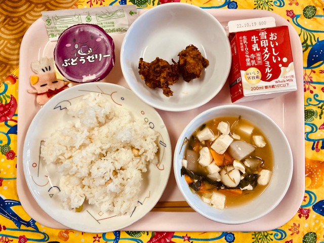 ピンクのトレーに給食が右上から反時計回りで牛乳、鶏肉の衣揚げ、ぶどうゼリー、コック帽をかぶった布製マスコット、まめまめごはん、月見汁と並べられている写真