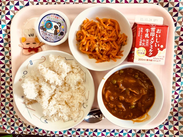 ピンクのトレーに給食が右上から反時計回りで牛乳、にんじんシリシリ、ブルーベリーゼリー、コック帽をかぶった布製マスコット、ごはん、ハヤシライスのルーと並べられている写真