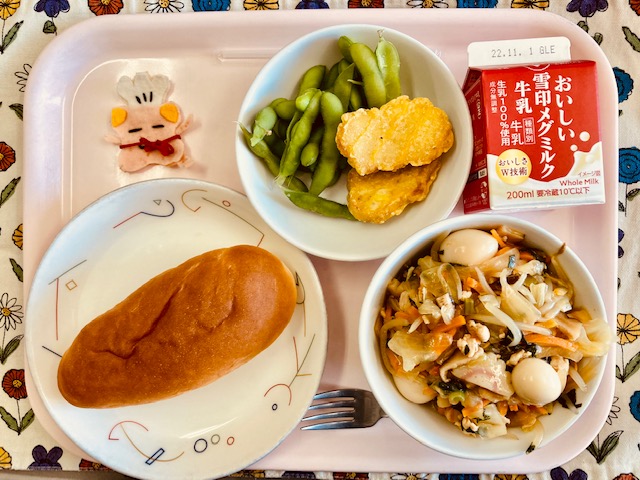 ピンクのトレーに給食が右上から反時計回りで牛乳、おさかなナゲット、枝豆、コック帽をかぶった布製マスコット、あじつけパン、やきそばあんかけと並べられている写真