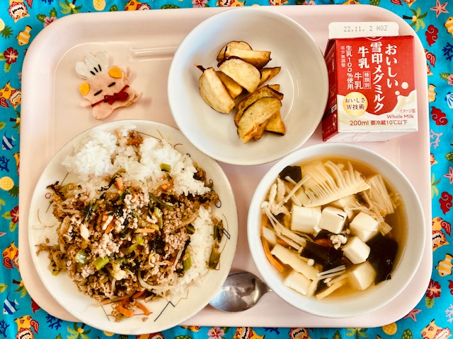 ピンクのトレーに給食が右上から反時計回りで牛乳、さつまいものからあげ、コック帽をかぶった布製マスコット、ビビンバ、わかめスープと並べられている写真