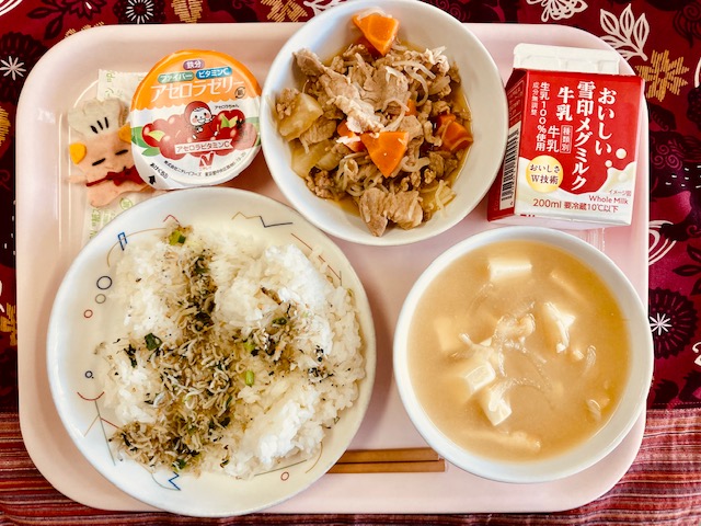 ピンクのトレーに給食が右上から反時計回りで牛乳、豚大根、アセロラゼリー、コック帽をかぶった布製マスコット、じゃこふりかけのごはん、みそ汁と並べられている写真