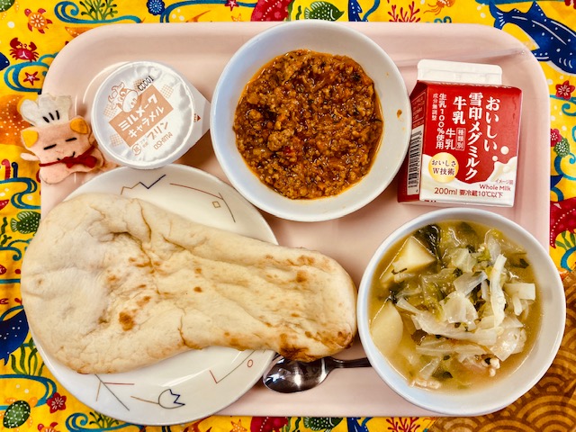 ピンクのトレーに給食が右上から反時計回りで牛乳、キーマカレー、ミルメークキャラメルプリン、コック帽をかぶった布製マスコット、ナン、やさいのスープ煮と並べられている写真