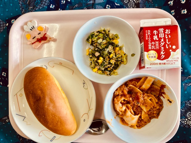 ピンクのトレーに給食が右上から反時計回りで牛乳、こまツナサラダ、コック帽をかぶった布製マスコット、パンプキンパン、ラザニアと並べられている写真