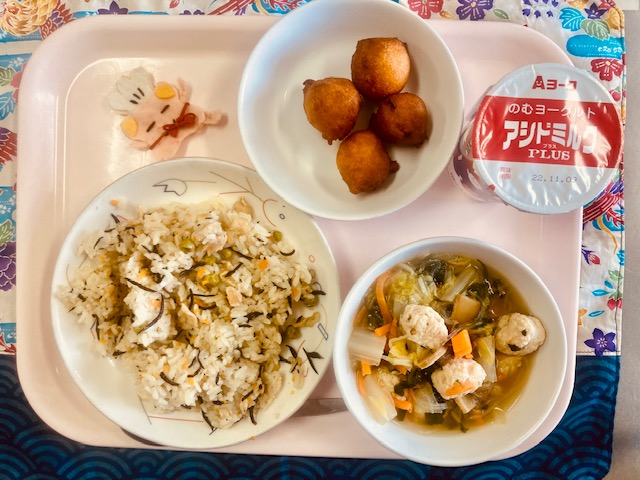 ピンクのトレーに給食が右上から反時計回りで飲むヨーグルト、パンプキンボール、コック帽をかぶった布製マスコット、ひじきピラフ、鶏団子と白菜のスープと並べられている写真