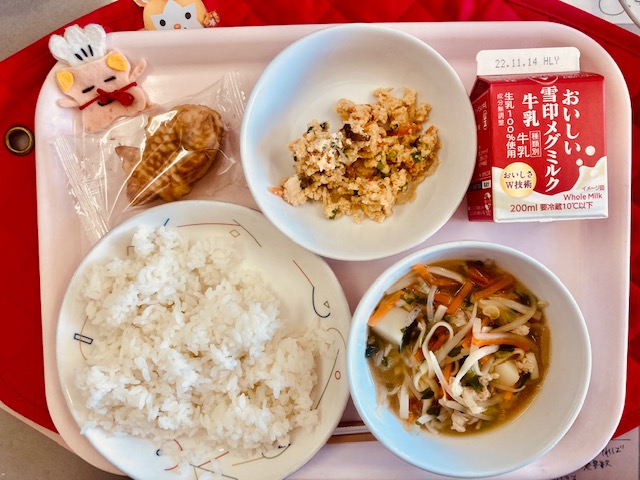 ピンクのトレーに給食が右上から反時計回りで牛乳、卯の花丼の具、コック帽をかぶった布製マスコット、プチたいやき、ごはん、あんかけ汁と並んでいる写真