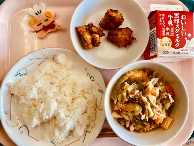 ピンクのトレーに給食が右上から反時計回りで牛乳、さばのカレー揚げ、コック帽をかぶった布製マスコット、ごはん、ジャージャン豆腐と並んでいる写真