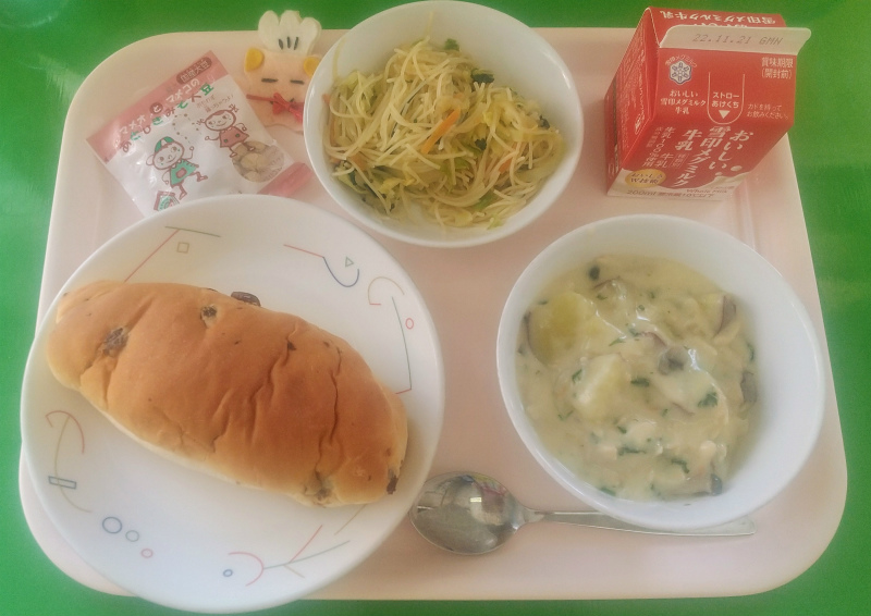 ピンクのトレーに給食が右上から反時計回りで牛乳、ビーフンソテー、コック帽をかぶった布製マスコット、味噌大豆、ぶどうパン、秋の味覚シチューと並んでいる写真