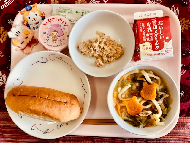 ピンクのトレーに給食が右上から反時計回りで牛乳、ツナ、いちごプリン、ユニフォーム姿のマスコット、コック帽をかぶった布製マスコット、パン、肉ネギうどんと並んでいる写真