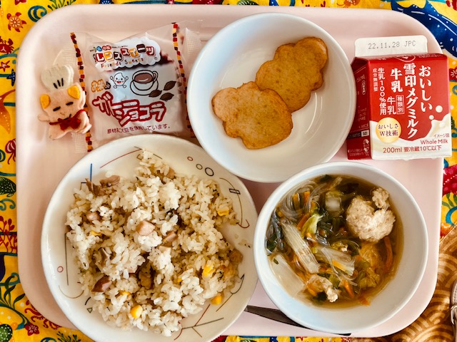 ピンクのトレーに給食が右上から反時計回りで牛乳、木の葉のさつま揚げ、米粉のガトーショコラ、コック帽をかぶった布製マスコット、キノコとベーコンのピラフ、白菜と肉団子のスープと並んでいる写真