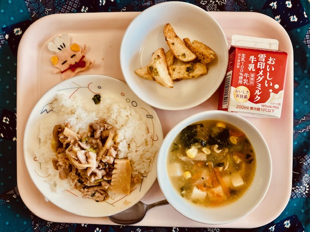 ピンクのトレーに給食が右上から反時計回りで牛乳、フライドポテト、コック帽をかぶった布製マスコット、肉あんかけごはん、コーン中華スープと並んでいる写真