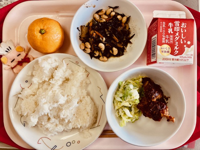 ピンクのトレーに給食が右上から反時計回りで牛乳、ひじきのいり煮、みかん、コック帽をかぶった布製マスコット、ごはん、キャベツソテー、みそかつと並んでいる写真