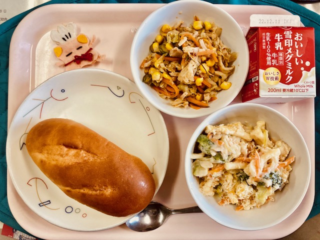 ピンクのトレーに給食が右上から反時計回りで牛乳、サラダでげんき、コック帽をかぶった布製マスコット、ミルクパン、とり肉とブロッコリーのグラタンと並んでいる写真