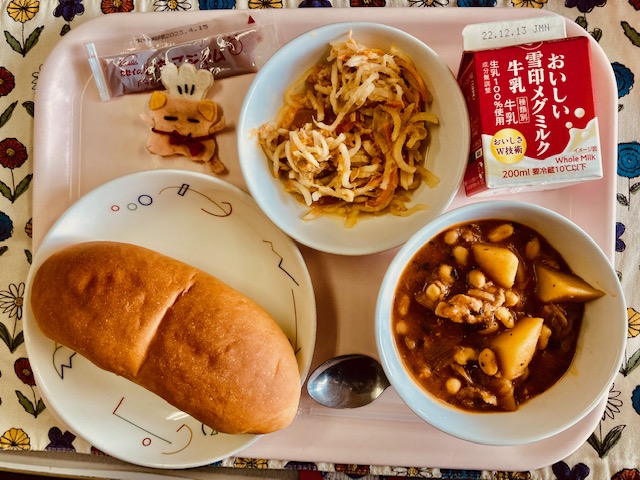 ピンクのトレーに給食が右上から反時計回りで牛乳、だいこんサラダ、いちごジャム、コック帽をかぶった布製マスコット、ソフトフランスパン、まめストロガノフと並んでいる写真