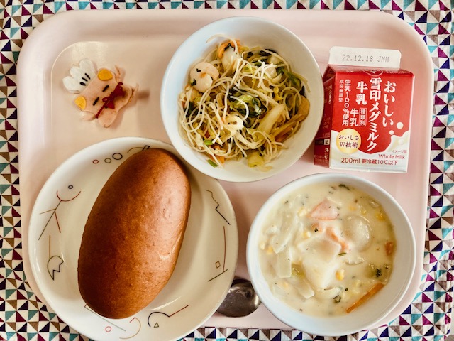 ピンクのトレーに給食が右上から反時計回りで牛乳、ビーフンソテー、コック帽をかぶった布製マスコット、くろパン、白菜とコーンのクリームシチューと並んでいる写真