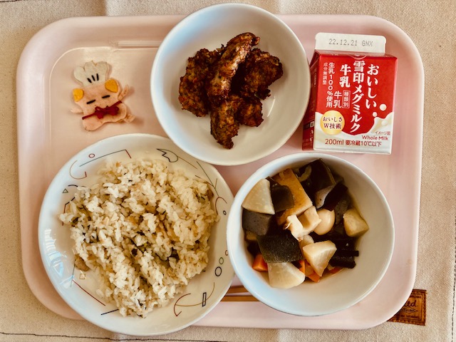 ピンクのトレーに給食が右上から反時計回りで牛乳、豚肉の風味揚げ、コック帽をかぶった布製マスコット、とりなめし、おでんと並んでいる写真