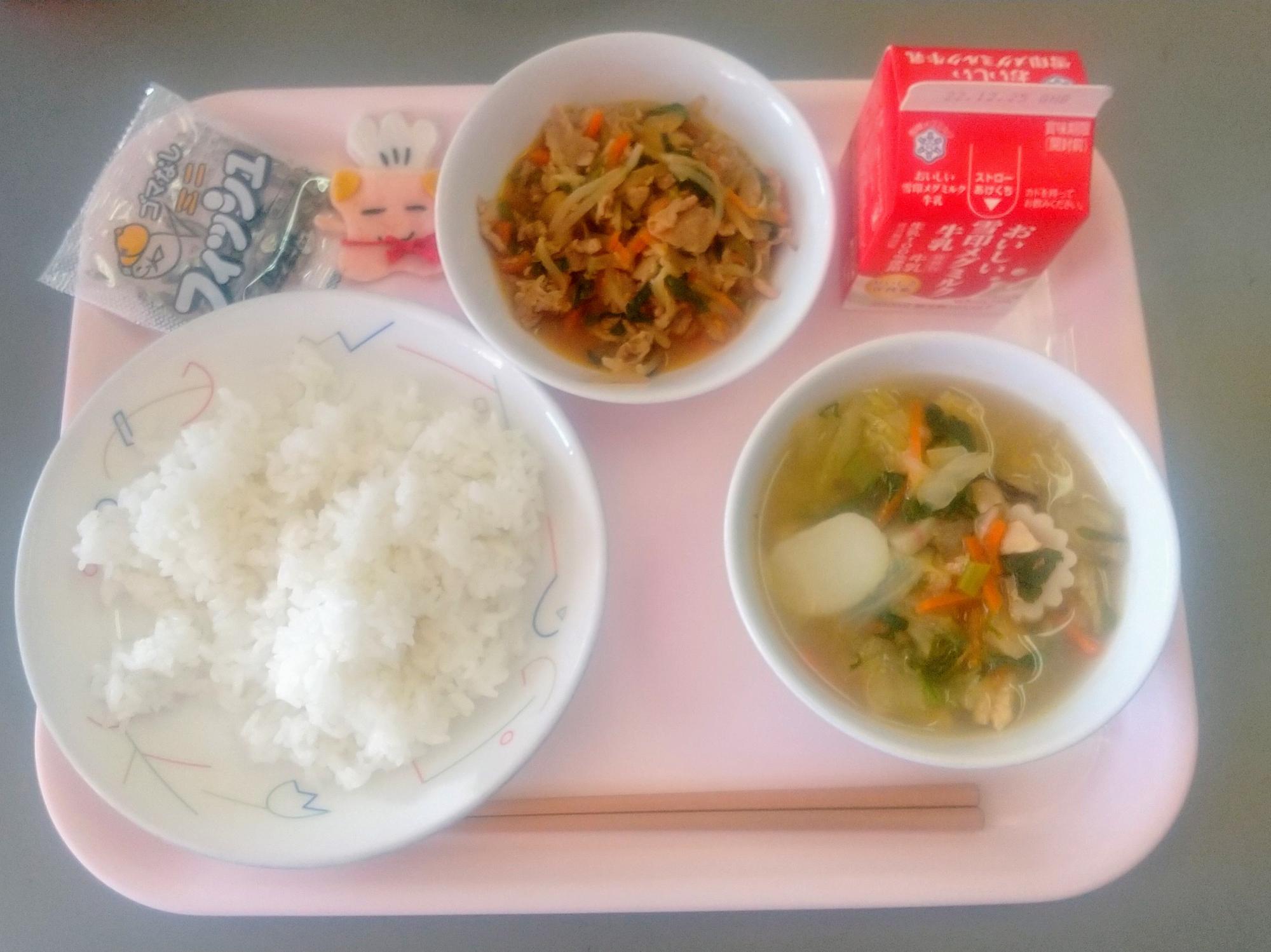 ピンクのトレーに給食が右上から反時計回りで牛乳、豚キムチ、コック帽をかぶった布製マスコット、こざかな、ごはん、トックスープと並んでいる写真
