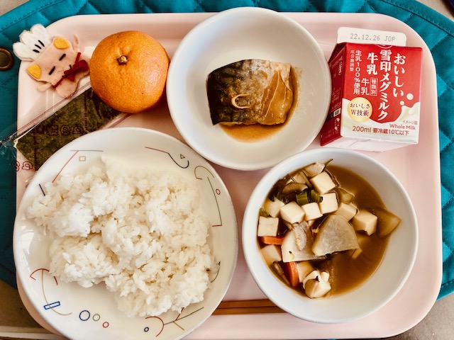 ピンクのトレーに給食が右上から反時計回りで牛乳、さばのみそに、みかん、コック帽をかぶった布製マスコット、のり、ごはん、けんちんじると並んでいる写真