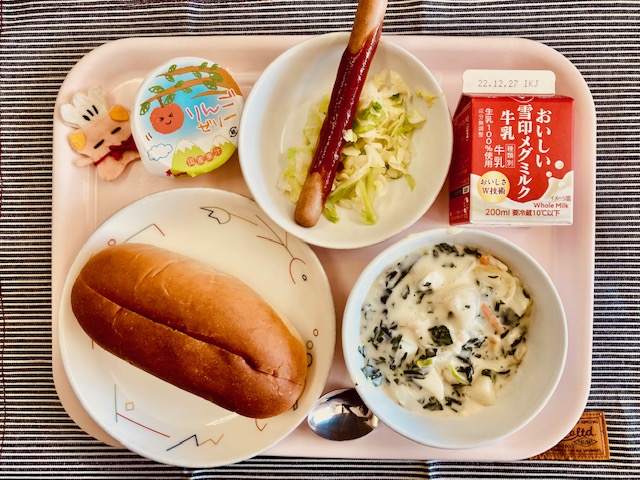 ピンクのトレーに給食が右上から反時計回りで牛乳、野菜、ウインナー、りんごゼリー、コック帽をかぶった布製マスコット、パン、ほうれん草のミルクスープと並んでいる写真