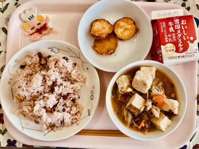 ピンクのトレーに給食が右上から反時計回りで牛乳、さつまいもの天ぷら、コック帽をかぶった布製マスコット、しあわせごはん、すきやきと並べられている写真