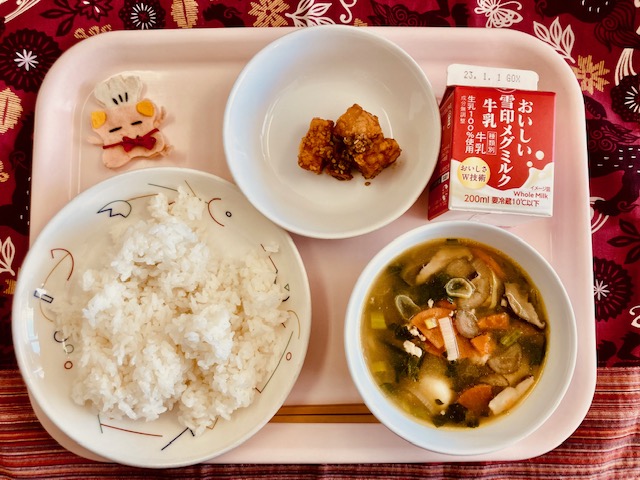 ピンクのトレーに給食が右上から反時計回りで牛乳、かじきのあずま煮、コック帽をかぶった布製マスコット、ごはん、芋もち汁と並べられている写真