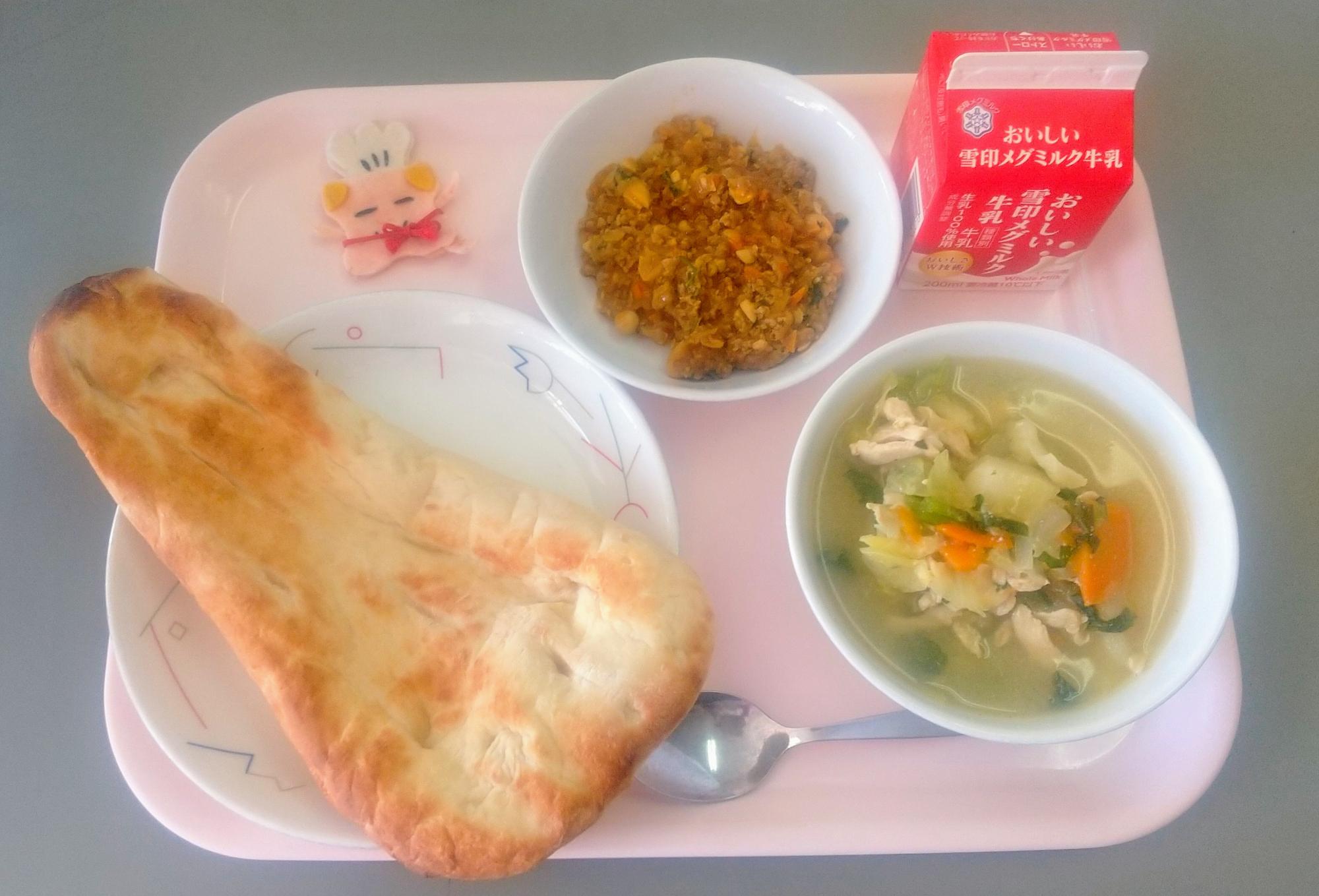ピンクのトレーに給食が右上から反時計回りで牛乳、ドライカレー、コック帽をかぶった布製マスコット、ナン、やさいのスープ煮と並べられている写真