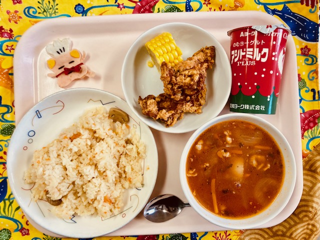 ピンクのトレーに給食が右上から反時計回りで発酵乳、コーン、フライドチキン、コック帽をかぶった布製マスコット、ガーリックピラフ、ミネストローネと並べられている写真