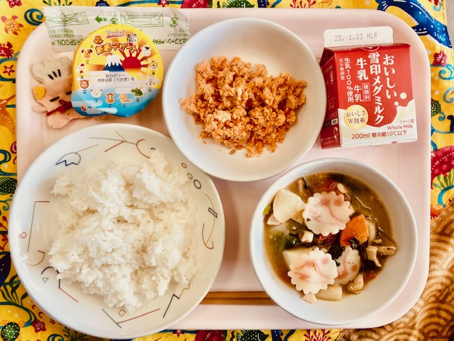 ピンクのトレーに給食が右上から反時計回りで牛乳、さけそぼろごはん、デザート、コック帽をかぶった布製マスコット、ごはん、白玉雑煮と並べられている写真