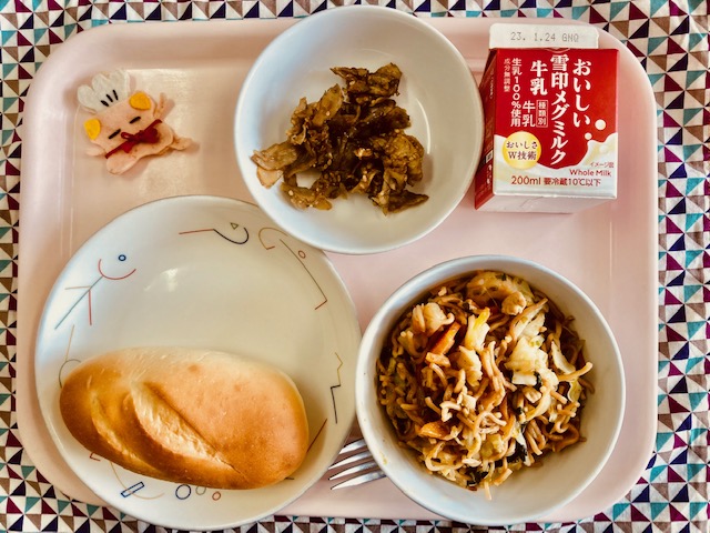 ピンクのトレーに給食が右上から反時計回りで牛乳、ごぼうのあまからだれからめ、コック帽をかぶった布製マスコット、ミルクパン、ソース焼きそばと並べられている写真