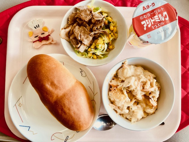 ピンクのトレーに給食が右上から反時計回りでのむヨーグルト、豚肉の和風マリネ野菜添え、コック帽をかぶった布製マスコット、ソフトフランスパン、ポテトグラタンと並べられている写真