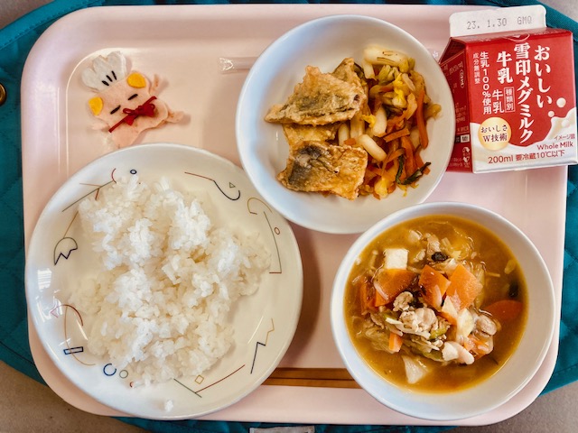 ピンクのトレーに給食が右上から反時計回りで牛乳、白菜漬け、あじの唐揚げ、コック帽をかぶった布製マスコット、ごはん、すきやきと並べられている写真