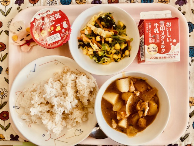 ピンクのトレーに給食が右上から反時計回りで牛乳、やさいソテー、ヨーグルト、コック帽をかぶった布製マスコット、ごはん、チキンカレーのルーと並べられている写真