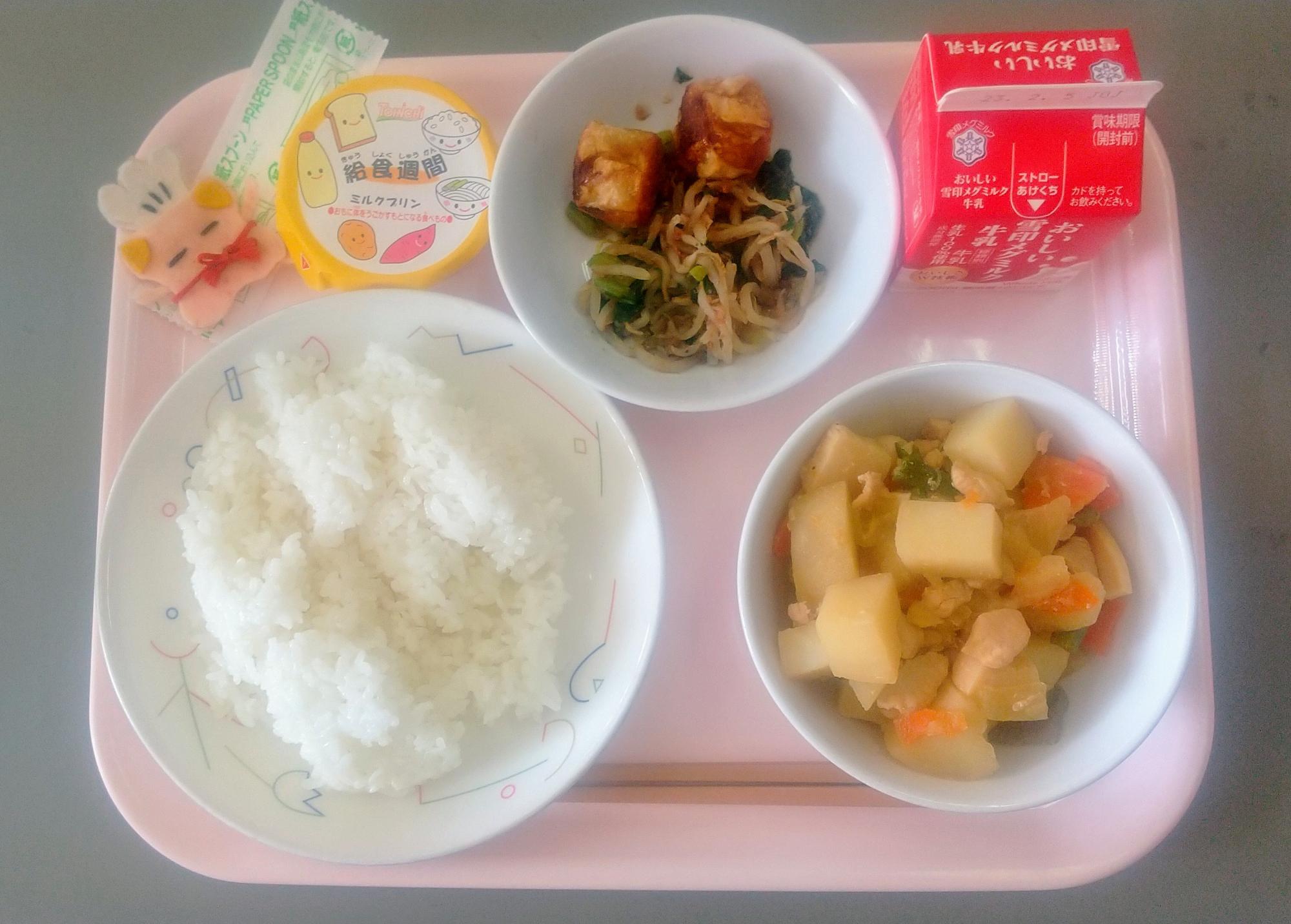 ピンクのトレーに給食が右上から反時計回りで牛乳、もやいsのおかかあえ、揚げシューマイ、デザート、コック帽をかぶった布製マスコット、ごはん、やさいのうま煮と並べられている写真