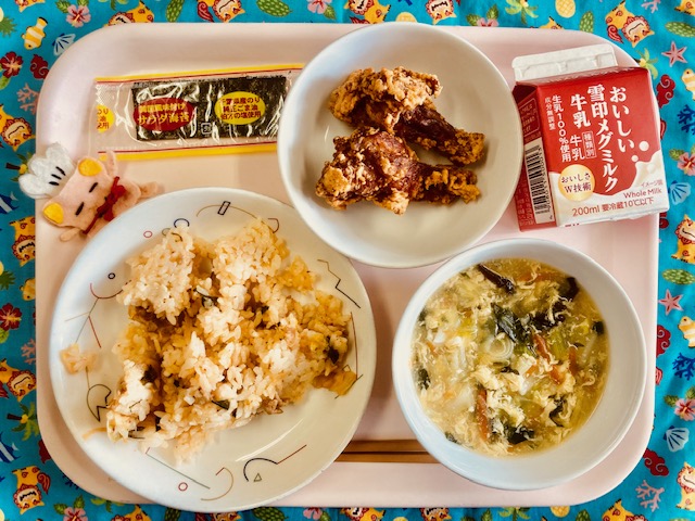 ピンクのトレーに給食が右上から反時計回りで牛乳、フライドチキン、サラダのり、コック帽をかぶった布製マスコット、キムチごはん、キャベツとたまごのスープと並べられている写真