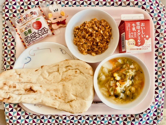 ピンクのトレーに給食が右上から反時計回りで牛乳、ドライカレー、コック帽をかぶった布製マスコット、国産りんごのタルト、ナン、やさいスープと並べられている写真