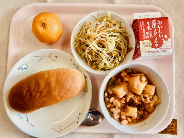 ピンクのトレーに給食が右上から反時計回りで牛乳、ビーフンソテー、ポンカン、あじつけパン、ポークビーンズと並べられている写真