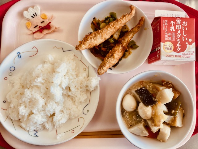 ピンクのトレーに給食が右上から反時計回りで牛乳、おひたし、ししゃもフライ、コック帽をかぶった布製マスコット、ごはん、にくみそおでんと並べられている写真