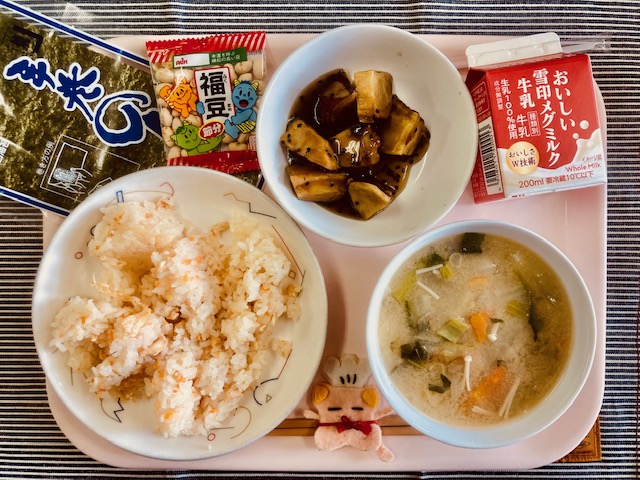 ピンクのトレーに給食が右上から反時計回りで牛乳、大学いも、福豆、のり、五目すし、コック帽をかぶった布製マスコット、いわしのつみれ汁と並べられている写真