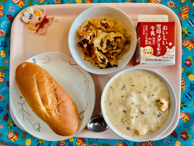 ピンクのトレーに給食が右上から反時計回りで牛乳、ハリハリサラダ、コック帽をかぶった布製マスコット、ミルクパン、オレンジボールシチューと並べられている写真