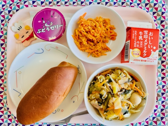 ピンクのトレーに給食が右上から反時計回りで牛乳、にんじんシリシリ、ぶどうゼリー、コック帽をかぶった布製マスコット、くろパン、やきそばあんかけと並べられている写真