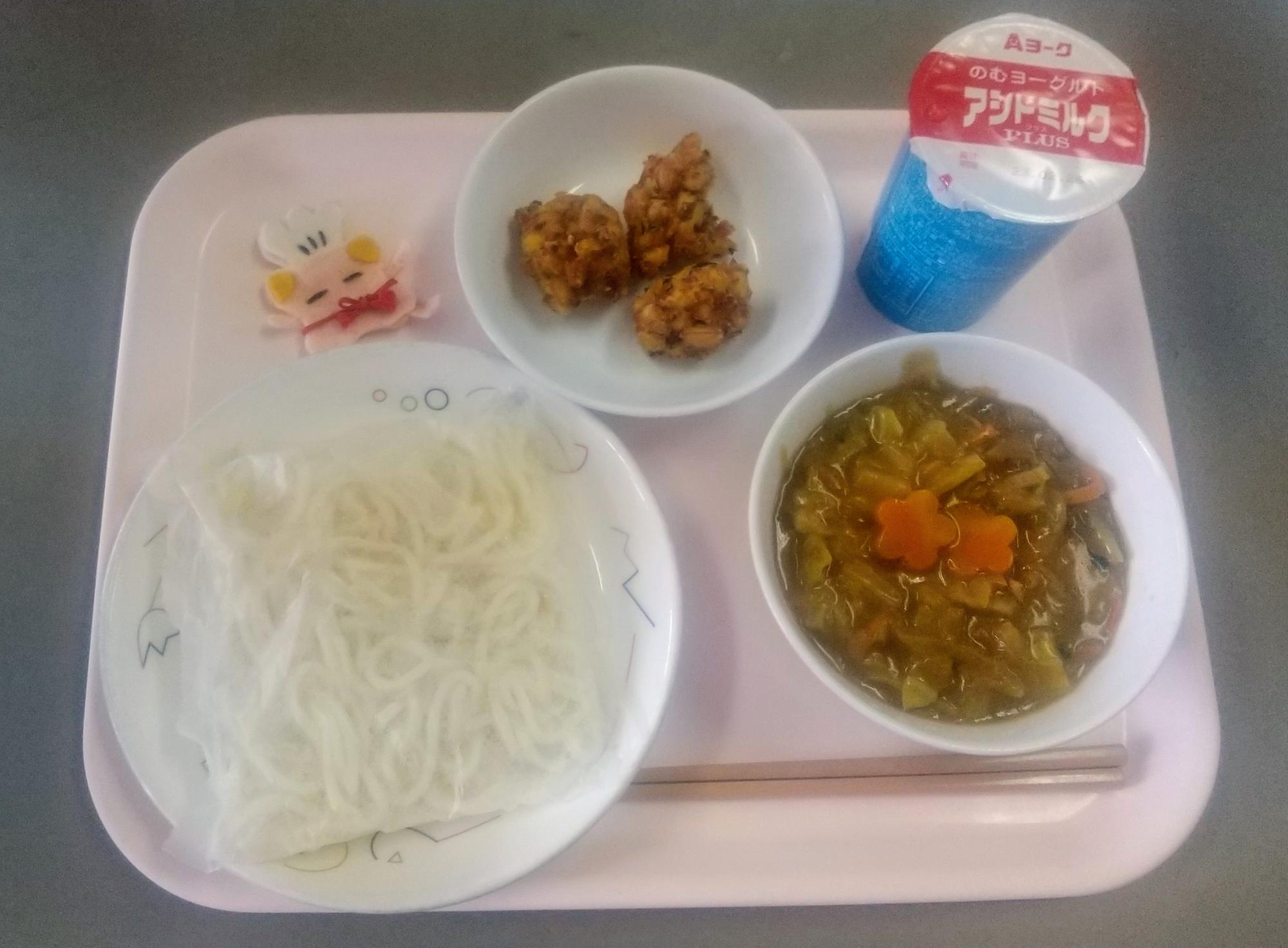 ピンクのトレーに給食が右上から反時計回りで発酵乳、がんせきあげ、コック帽をかぶった布製マスコット、ソフトめん、カレーあんかけのあんと並んでいる写真