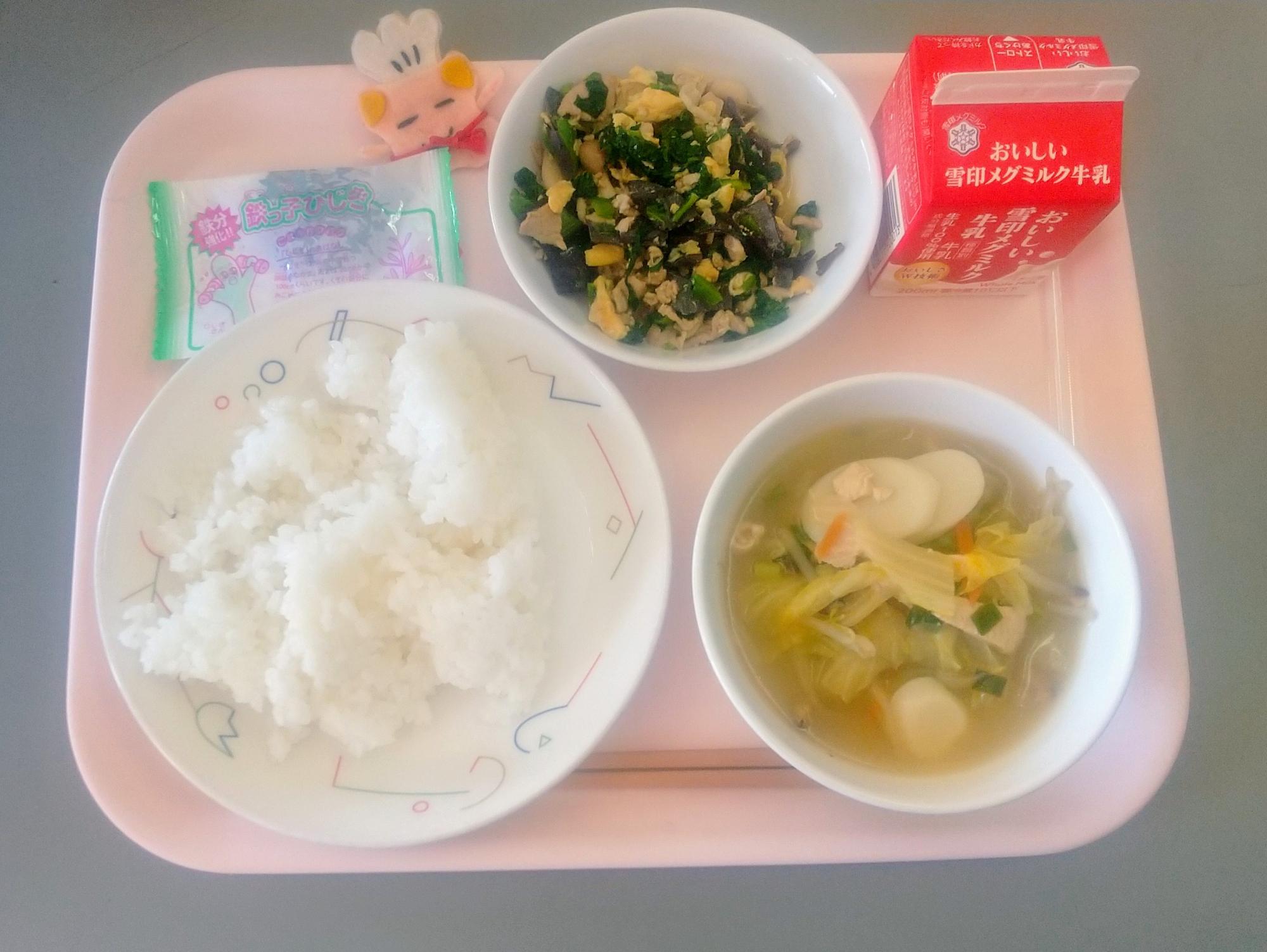ピンクのトレーに給食が右上から反時計回りで牛乳、小松菜と卵のオイスターソース炒め、コック帽をかぶった布製マスコット、佃煮、ごはん、トックスープと並んでいる写真