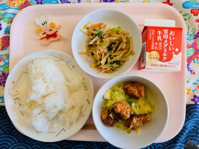 ピンクのトレーに給食が右上から反時計回りで牛乳、五目きんぴら、コック帽をかぶった布製マスコット、ごはん、キャベツソテー、めかじきフライと並んでいる写真