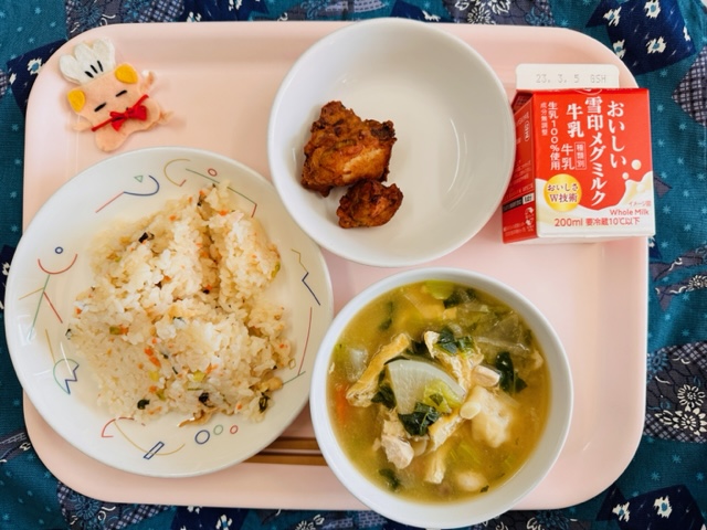 ピンクのトレーに給食が右上から反時計回りで牛乳、さわらのかおり揚げ、コック帽をかぶった布製マスコット、かやくごはん、すいとんと並んでいる写真