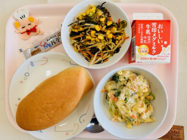 ピンクのトレーに給食が右上から反時計回りで牛乳、海藻サラダ、コック帽をかぶった布製マスコット、キャラメルパテ、コッペパン、マカロニの豆乳グラタンと並んでいる写真