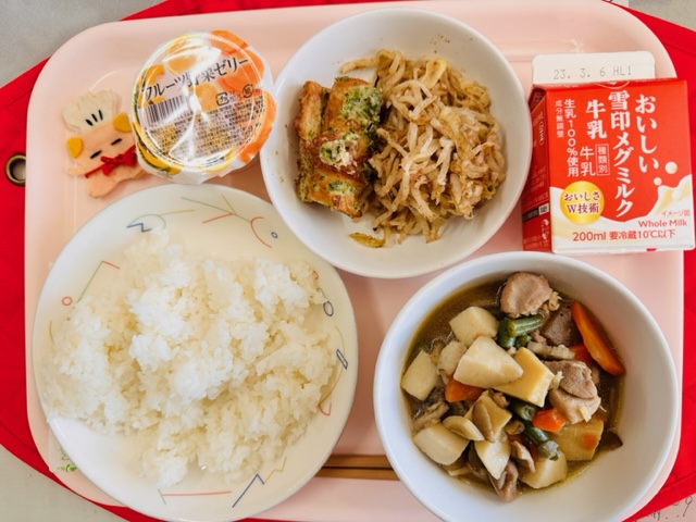 ピンクのトレーに給食が右上から反時計回りで牛乳、もやしのおかか和え、ちくわのみなと揚げ、フルーツゼリー、コック帽をかぶった布製マスコット、ごはん、筑前煮と並んでいる写真