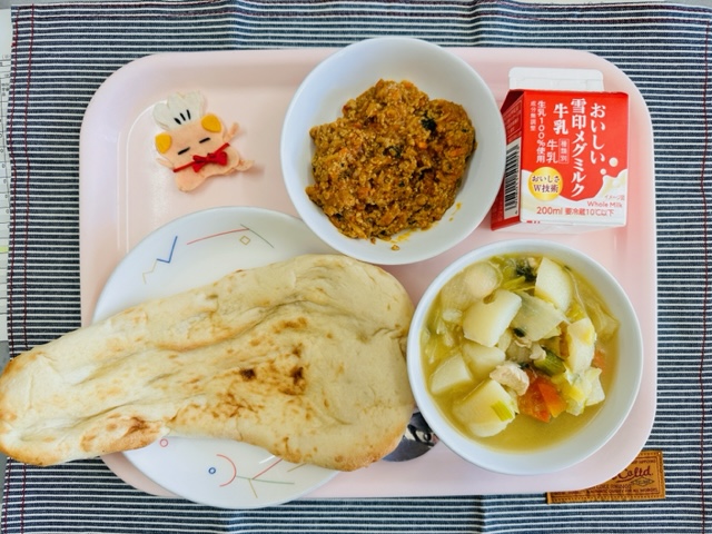 ピンクのトレーに給食が右上から反時計回りで牛乳、キーマカレー、コック帽をかぶった布製マスコット、ナン、やさいのスープ煮と並んでいる写真
