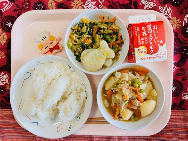 ピンクのトレーに給食が右上から反時計回りで牛乳、カラフルおひたしの上におさかなはんぺん、コック帽をかぶった布製マスコット、ごはん、にくじゃがと並んでいる写真